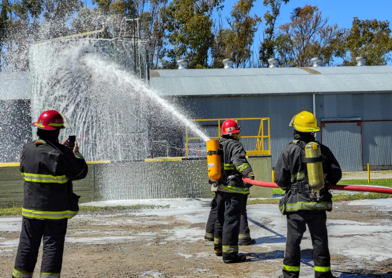 Bomberos entrenando con mangueras y equipos en un simulador al aire libre.