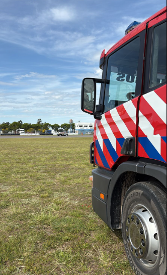 Vista lateral de un camión de bomberos con diseño a rayas rojas y blancas, en un área abierta con cielo azul y nubes.