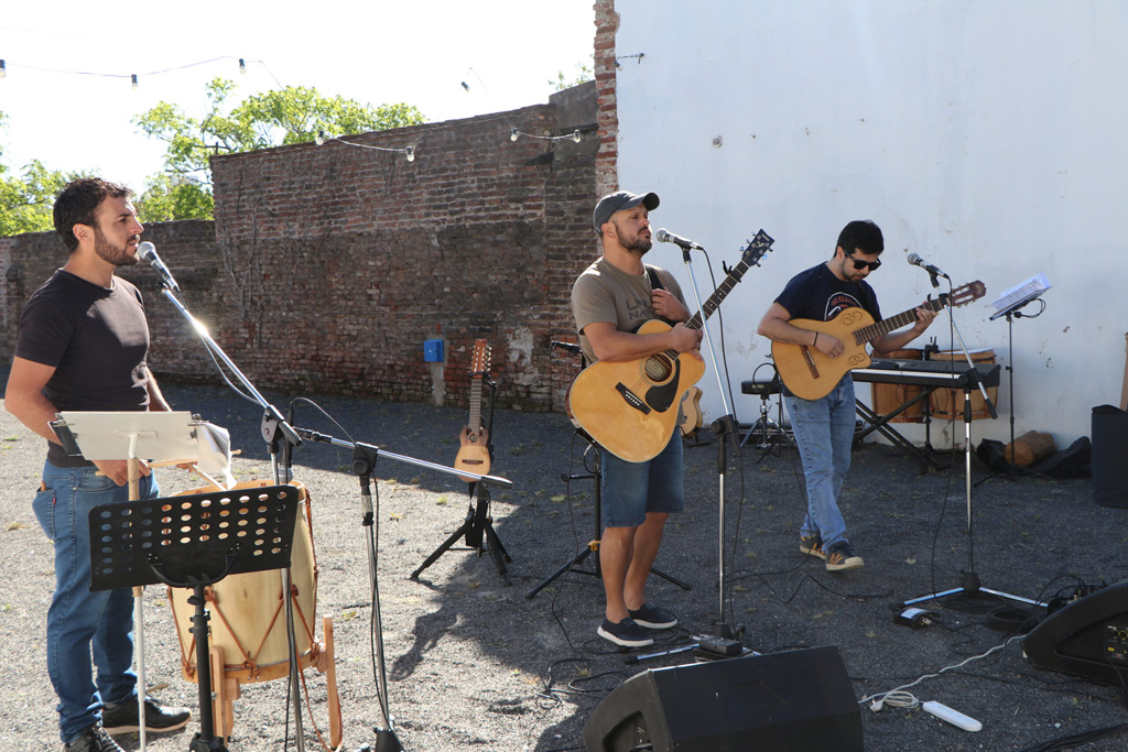 Tres músicos tocando y cantando en un escenario al aire libre, rodeados de un ambiente rústico.