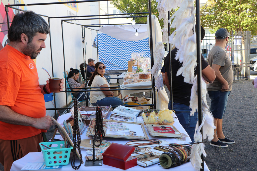 Un hombre con camiseta naranja sostiene una bebida mientras observa una mesa con joyería y artesanías en un mercado al aire libre, con otros visitantes al fondo.
