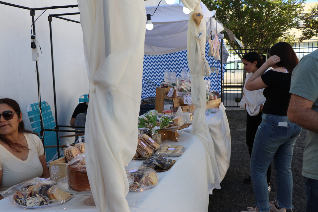 Una mujer sonríe mientras clientes exploran una mesa decorada con una variedad de dulces y postres en un mercado.