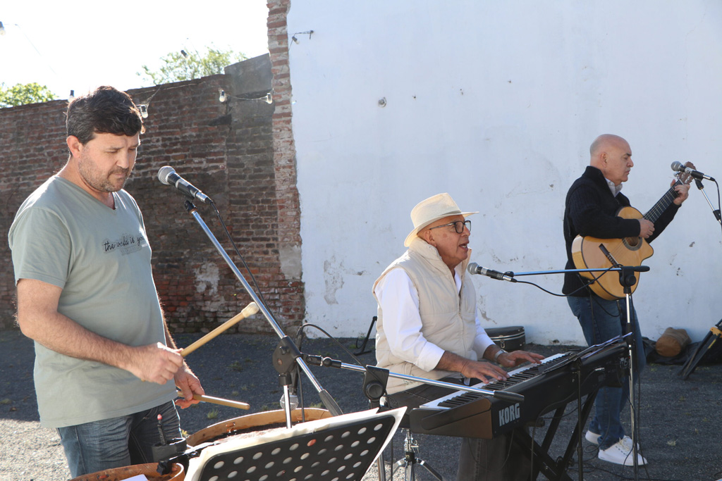 Tres músicos tocan en un escenario al aire libre, con un fondo de pared de ladrillo y un cielo claro.