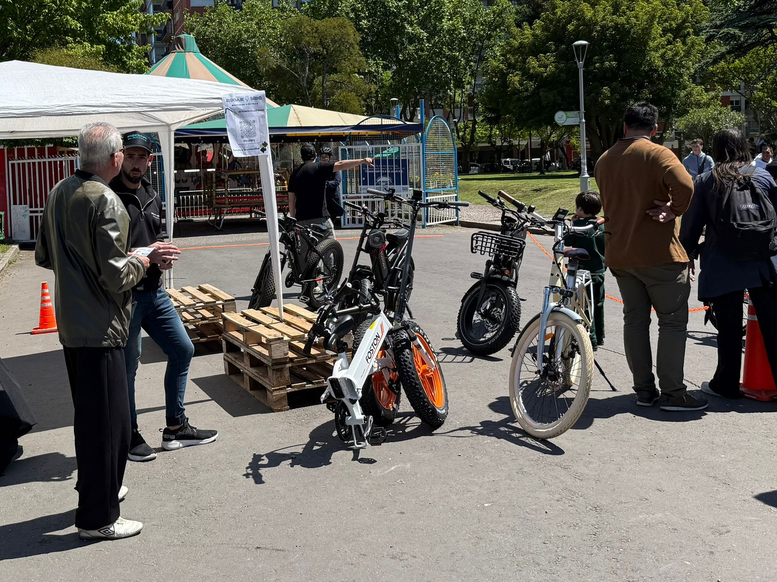 Personas conversando frente a un stand de bicicletas eléctricas en un parque, con bicicletas expuestas y un ambiente animado.